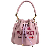 Borsa secchiello rosa in pelle con scritta "The Bucket", manico superiore, dettagli dorati, chiusura a coulisse. Accessorio moda elegante.