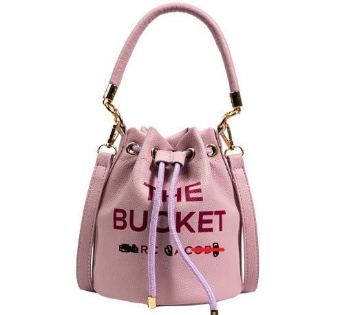 Borsa secchiello rosa in pelle con scritta "The Bucket", manico superiore, dettagli dorati, chiusura a coulisse. Accessorio moda elegante.