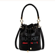 Borsa secchiello nera in pelle con manico e tracolla, dettagli dorati, scritta "The Bucket" e logo rosso. Elegante e moderna, perfetta per ogni occasione.