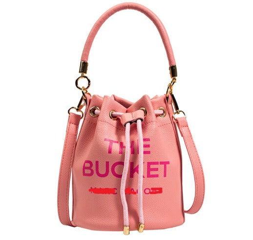Borsa a secchiello rosa in pelle sintetica con manico e tracolla, dettagli dorati, scritta "The Bucket". Accessorio moda donna elegante.