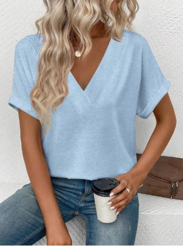 Donna con maglietta azzurra scollo a V, capelli biondi ondulati, jeans, tiene bicchiere di caffè. Moda casual femminile, abbigliamento estivo.