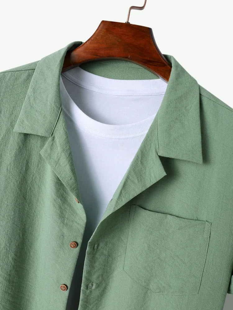 Camicia verde chiaro in lino con colletto classico, bottoni in legno e tasca frontale, su gruccia di legno. Sotto, t-shirt bianca a girocollo.