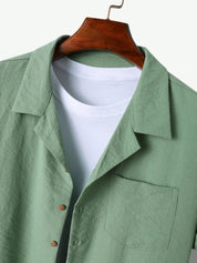 Camicia verde chiaro in lino con colletto classico, bottoni in legno e tasca frontale, su gruccia di legno. Sotto, t-shirt bianca a girocollo.