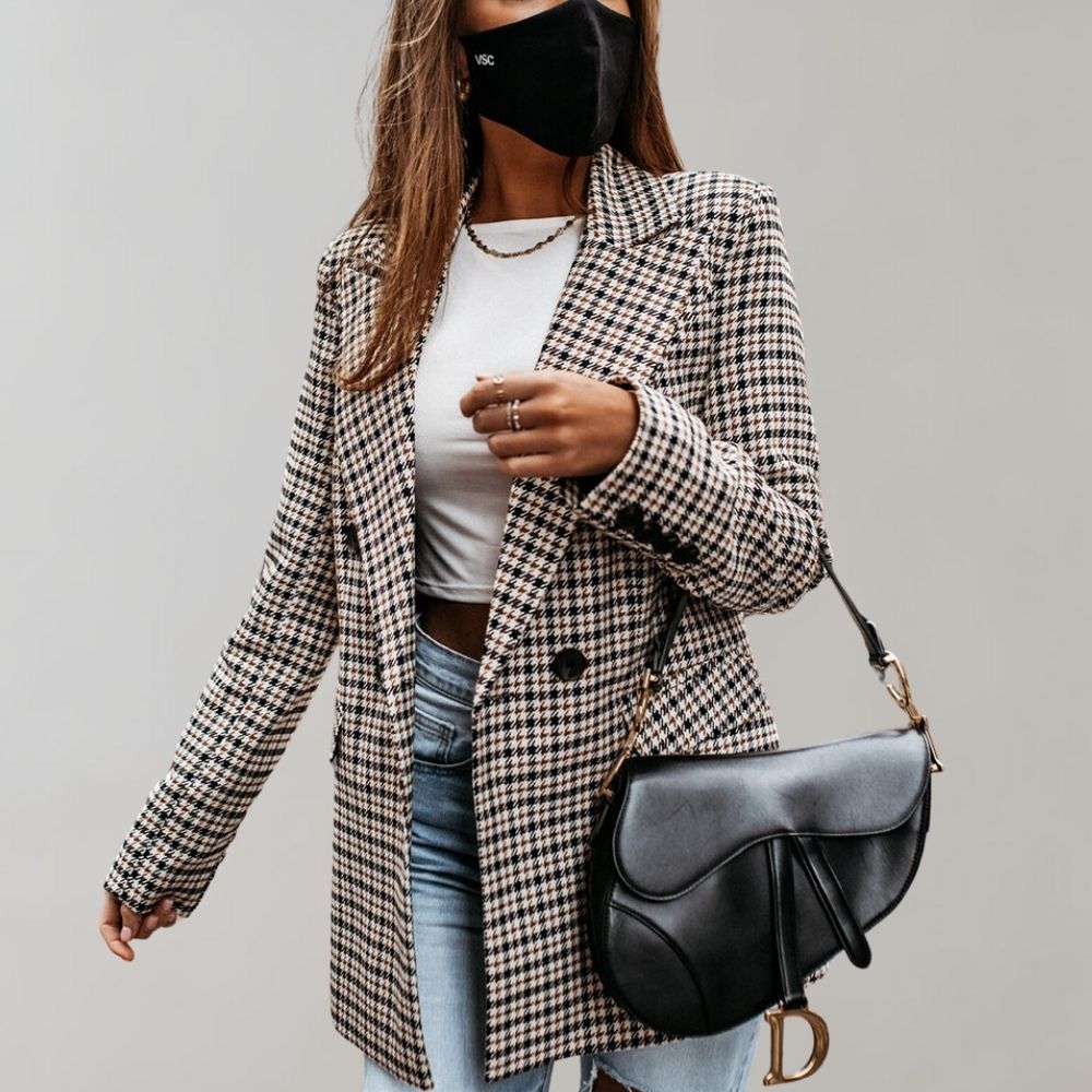 Donna con blazer a quadri, jeans e borsa a tracolla nera. Indossa mascherina nera e maglietta bianca. Moda autunnale elegante e casual.