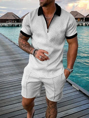 Uomo in completo estivo bianco a motivi geometrici, camicia con zip e pantaloncini, su passerella di legno vicino al mare. Moda estiva casual.