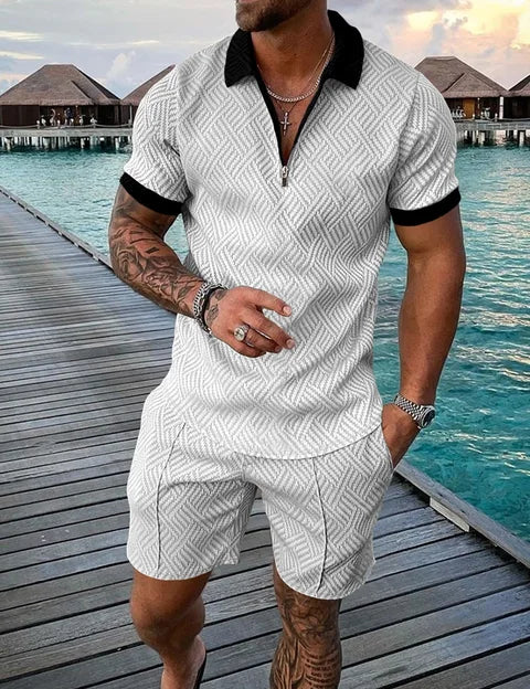 Uomo in completo estivo bianco a motivi geometrici, camicia con zip e pantaloncini, su passerella di legno vicino al mare. Moda estiva casual.