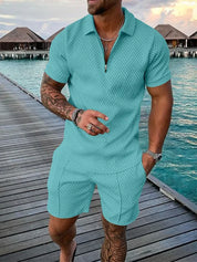 Uomo in completo estivo azzurro su passerella di legno, sfondo mare e bungalow. Moda maschile, abbigliamento casual, vacanza tropicale.