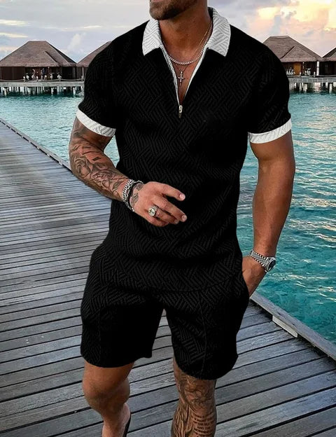 Uomo in completo estivo nero con motivo geometrico, camicia a maniche corte e pantaloncini, su passerella di legno vicino al mare. Moda maschile elegante.