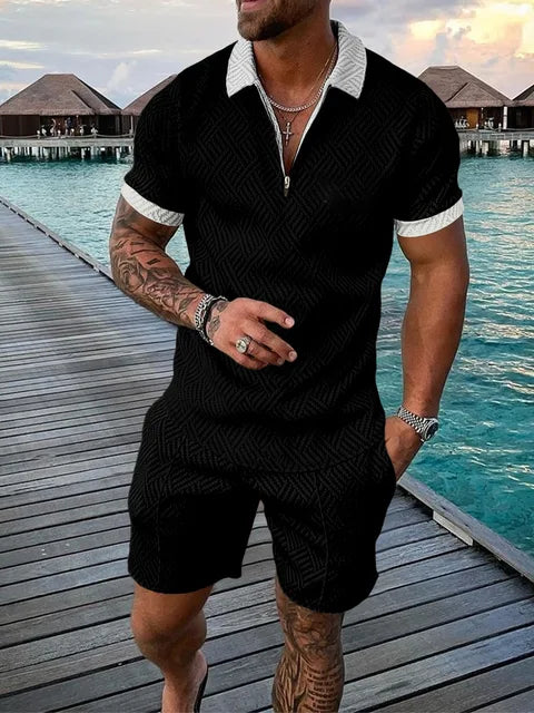Uomo in completo estivo nero con motivo geometrico, camicia a maniche corte e pantaloncini, su passerella di legno vicino al mare. Moda maschile elegante.