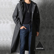 Cappotto impermeabile nero da uomo, lungo, con cappuccio, indossato su maglia nera e jeans. Stile casual, moda autunnale.