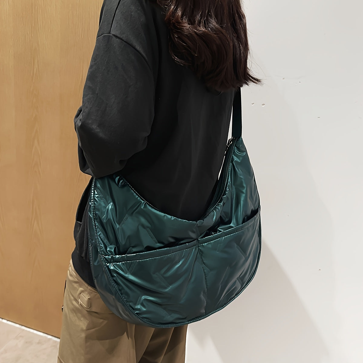 Donna con borsa a tracolla verde scuro in tessuto lucido, indossa maglia nera e pantaloni beige. Accessorio moda casual, stile urbano.