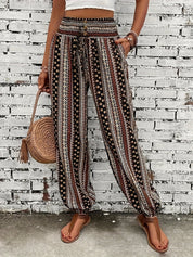 Pantaloni boho a righe etniche, donna, con borsa intrecciata e sandali, moda estiva casual, sfondo muro bianco.