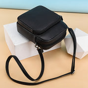Borsa a tracolla in pelle nera su sfondo beige e azzurro, design elegante e compatto, ideale per moda donna, accessori di tendenza.
