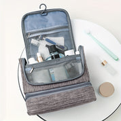 Beauty case da viaggio grigio aperto con gancio, scomparti a rete, contenente articoli da toeletta come spazzolino, flaconi e rasoio.