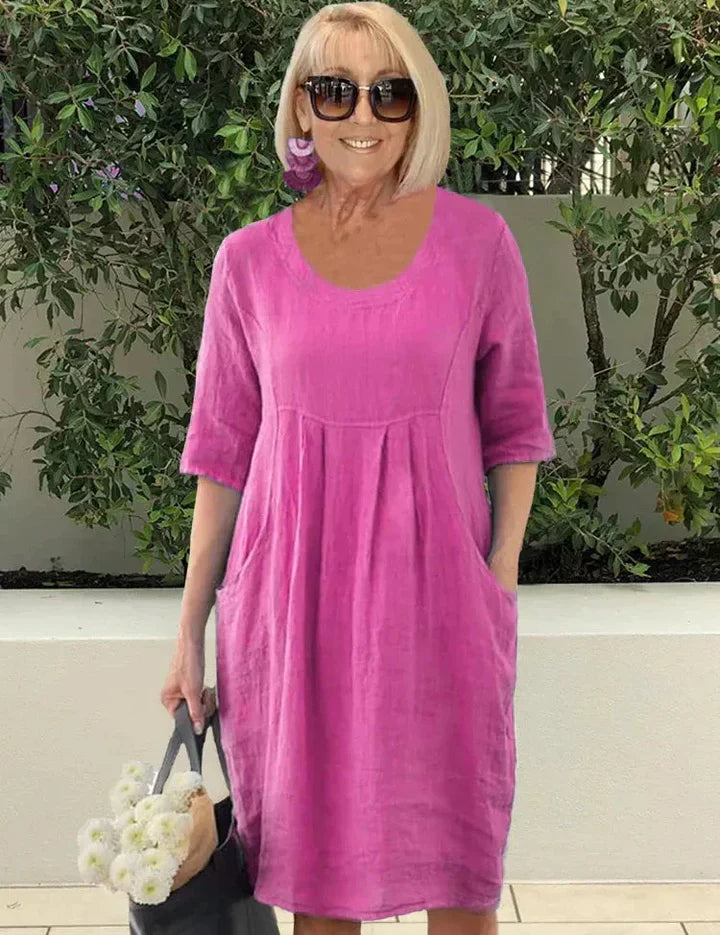 Donna con abito estivo rosa in lino, maniche corte, tasche laterali, occhiali da sole, orecchini viola, borsa nera con fiori bianchi.