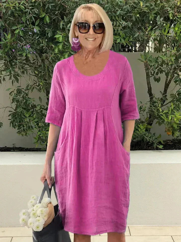Donna con abito estivo rosa in lino, maniche corte, tasche laterali, occhiali da sole, orecchini viola, borsa nera con fiori bianchi.