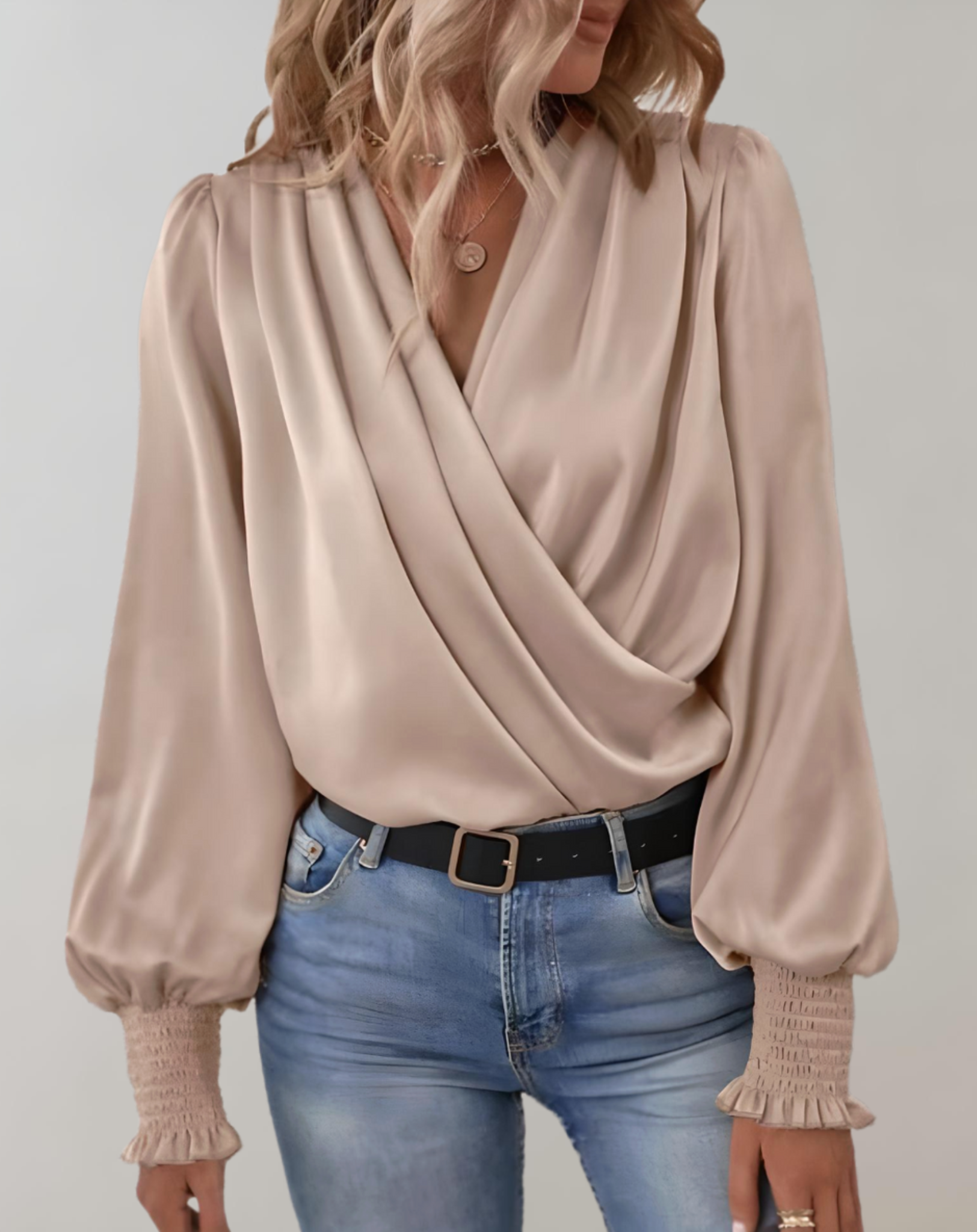 Blusa elegante in raso beige con scollo a V incrociato, maniche lunghe a sbuffo e polsini arricciati, abbinata a jeans chiari.