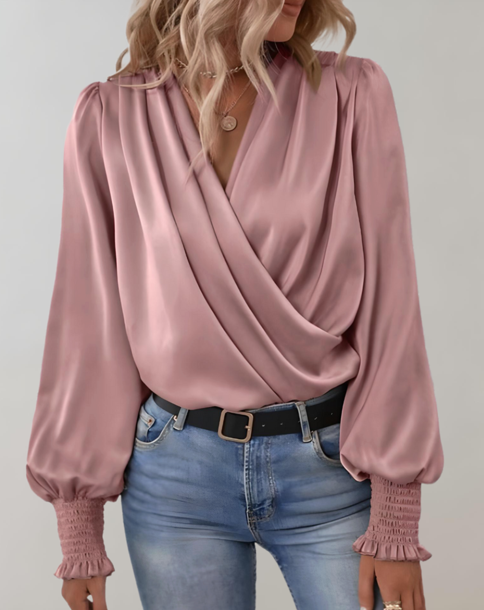 Blusa rosa in raso con scollo a V incrociato, maniche lunghe a sbuffo e polsini arricciati, abbinata a jeans chiari. Moda elegante donna.