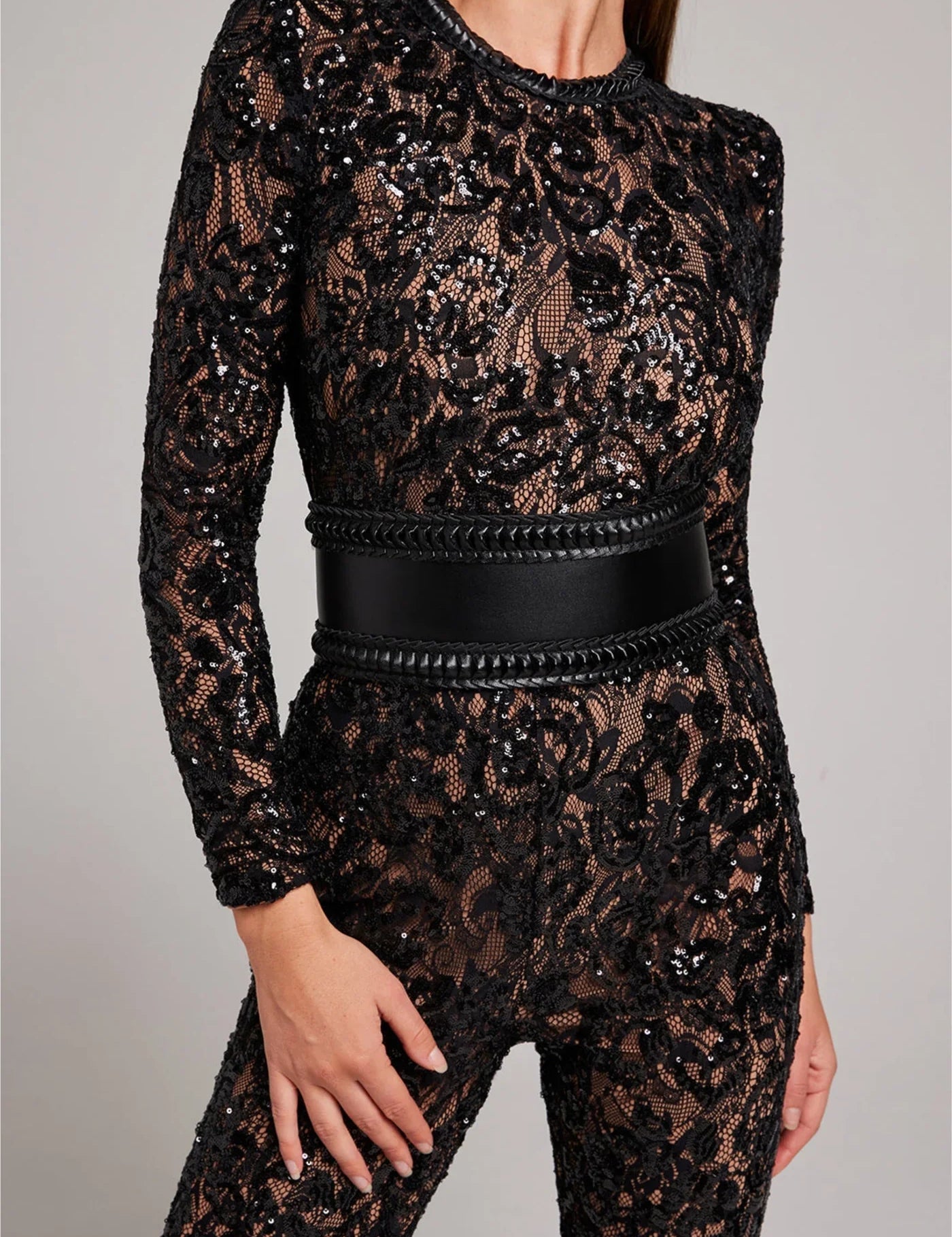 Tuta elegante in pizzo nero trasparente con dettagli floreali e cintura in pelle nera intrecciata. Abbigliamento da sera glamour e sofisticato.