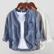 Camicia casual da uomo in lino, disponibile in blu, grigio e bianco, con colletto alla coreana e maniche lunghe, appesa su grucce di legno.