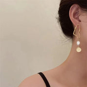 Orecchino pendente dorato con perla e moneta, design moderno, indossato su orecchio femminile. Gioielli eleganti, accessori moda donna.