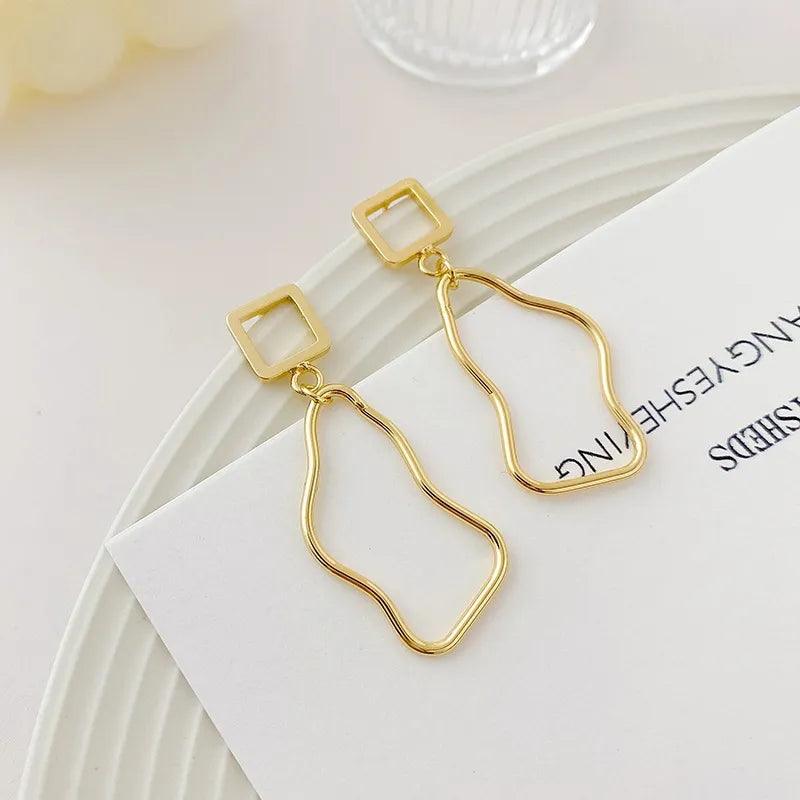 Orecchini pendenti dorati con design geometrico astratto su sfondo bianco. Accessori moda eleganti, ideali per look moderni e sofisticati.