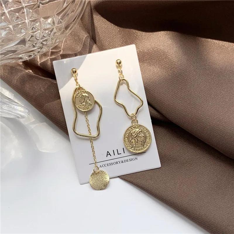 Orecchini pendenti asimmetrici in oro con design geometrico e monete, su sfondo marrone. Accessori moda eleganti per donna.