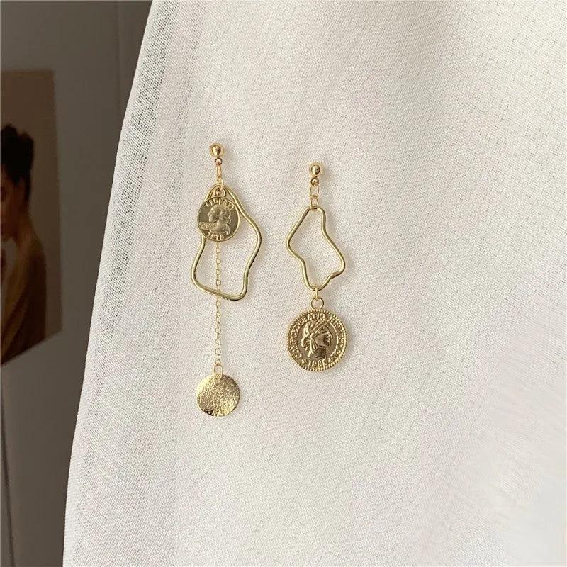Orecchini pendenti asimmetrici in oro, design geometrico con monete decorative, su sfondo bianco. Accessori moda eleganti e moderni.