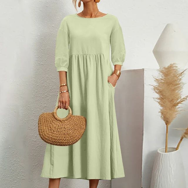 Abito lungo verde chiaro con maniche a tre quarti, tasche laterali, borsa in paglia intrecciata, sfondo bianco minimalista, moda donna elegante.