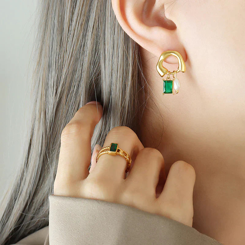 Orecchini pendenti in oro con pietra verde e perla, abbinati a un anello coordinato su mano femminile. Gioielli eleganti e moderni.