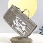 Astuccio grigio in tessuto con manico, tasca trasparente e chiusura a zip, posizionato su pietra. Design moderno, ideale per scuola o ufficio.