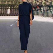 Tuta elegante blu scuro con mantella, donna in strada alberata. Abbigliamento moda femminile, stile sofisticato, tendenza autunno.