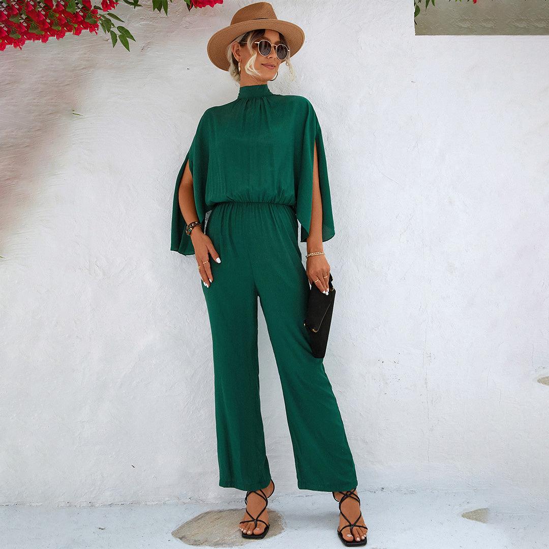 Donna in tuta verde elegante, cappello a tesa larga, occhiali da sole, sandali neri, borsa nera, sfondo bianco. Moda estiva, abbigliamento chic.