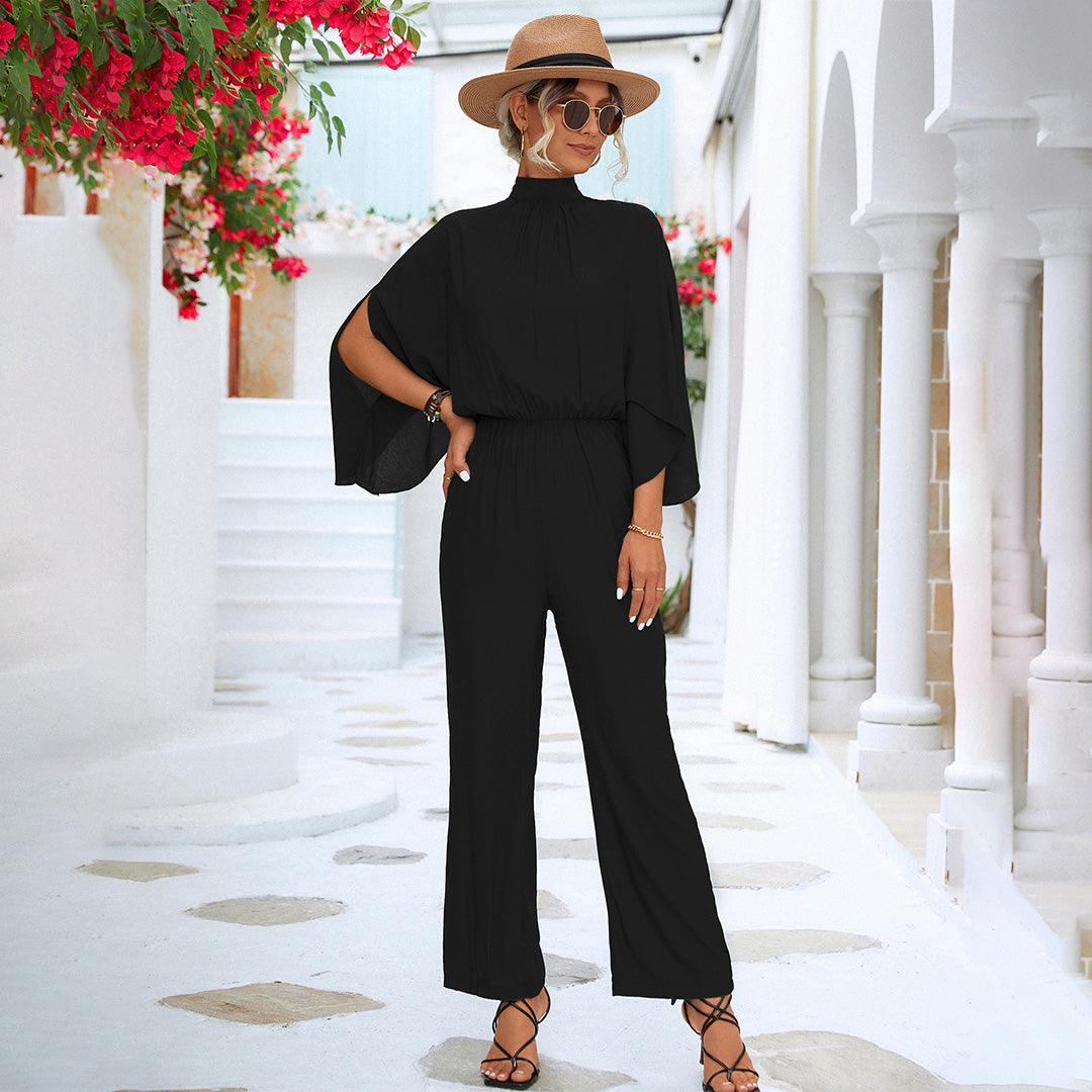 Donna con tuta nera elegante, cappello di paglia e occhiali da sole, in un ambiente mediterraneo con fiori rossi e colonne bianche.