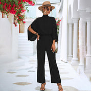Donna con tuta nera elegante, cappello di paglia e occhiali da sole, in un ambiente mediterraneo con fiori rossi e colonne bianche.