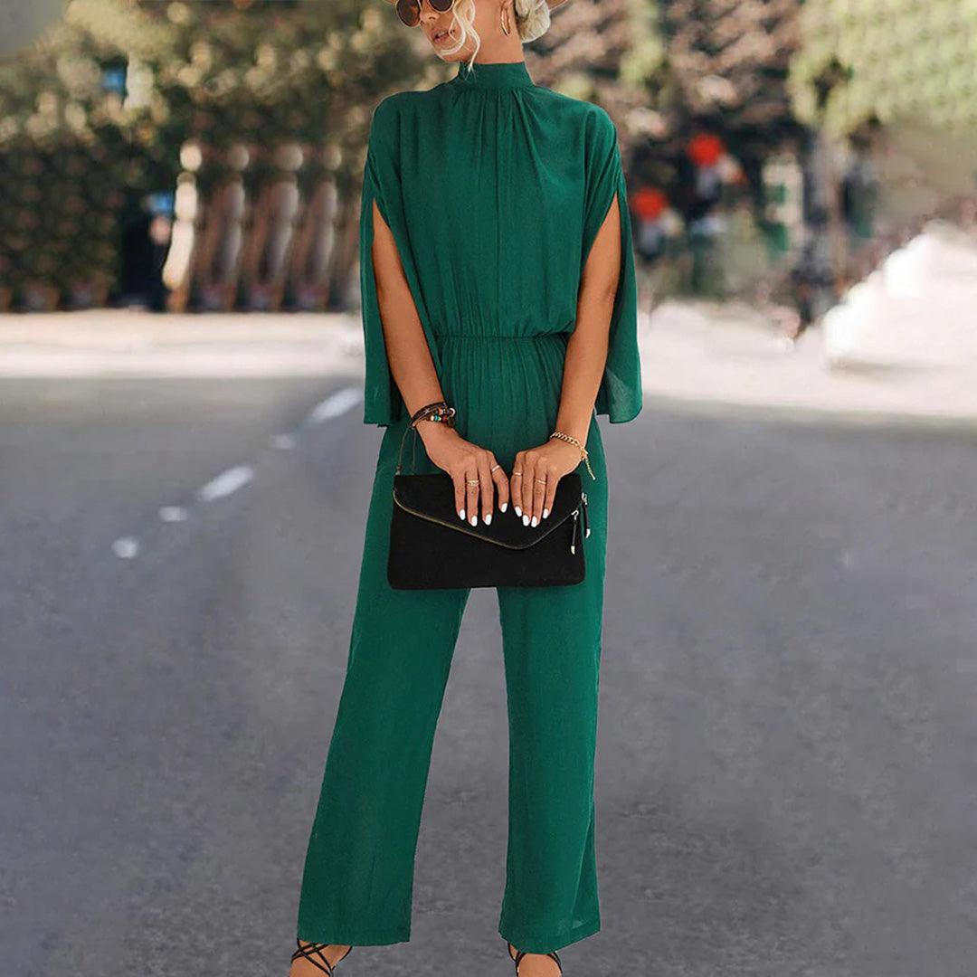 Donna in tuta verde elegante con maniche a mantella, occhiali da sole e borsa nera, su strada urbana. Moda femminile, abbigliamento chic.