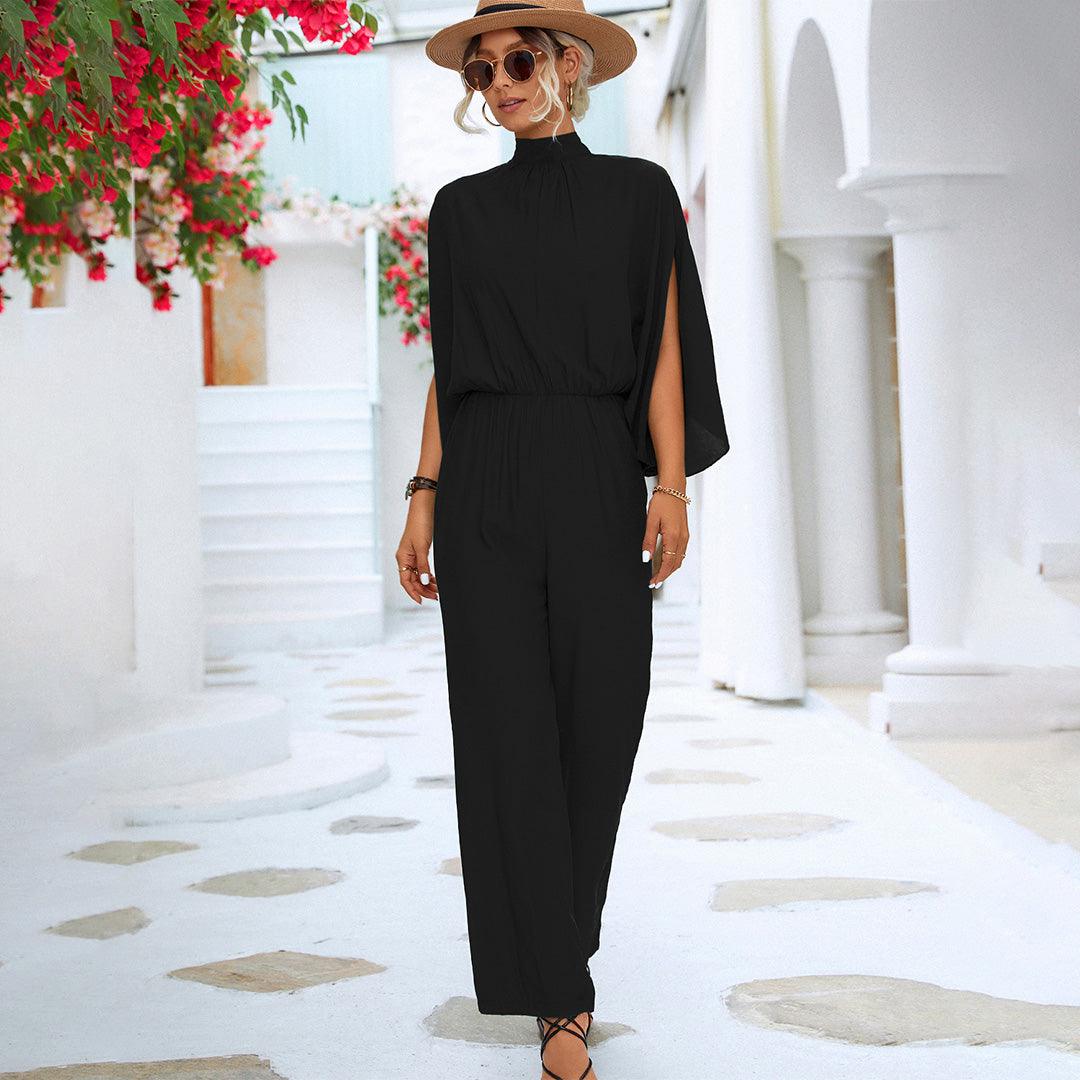 Donna con tuta nera elegante, cappello a tesa larga e occhiali da sole, cammina in strada bianca mediterranea con fiori rossi. Moda estiva chic.