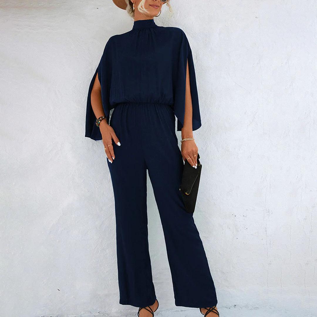 Tuta elegante blu scuro da donna con maniche a mantella, abbinata a borsa nera e sandali. Look sofisticato per eventi formali.