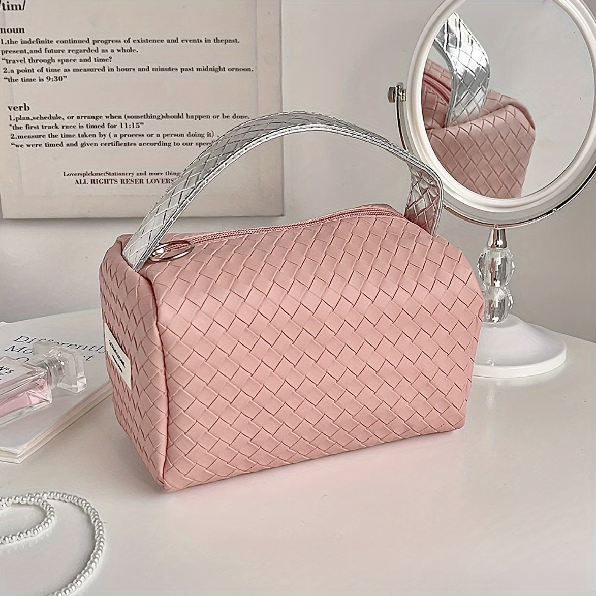 Borsa cosmetica rosa intrecciata su tavolo bianco con specchio rotondo. Elegante e pratica, ideale per organizzare trucchi e accessori di bellezza.