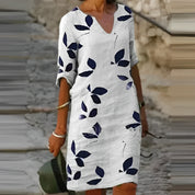 Abito estivo bianco con stampa foglie blu, maniche a tre quarti, scollo a V, indossato da donna su strada acciottolata. Moda casual elegante.