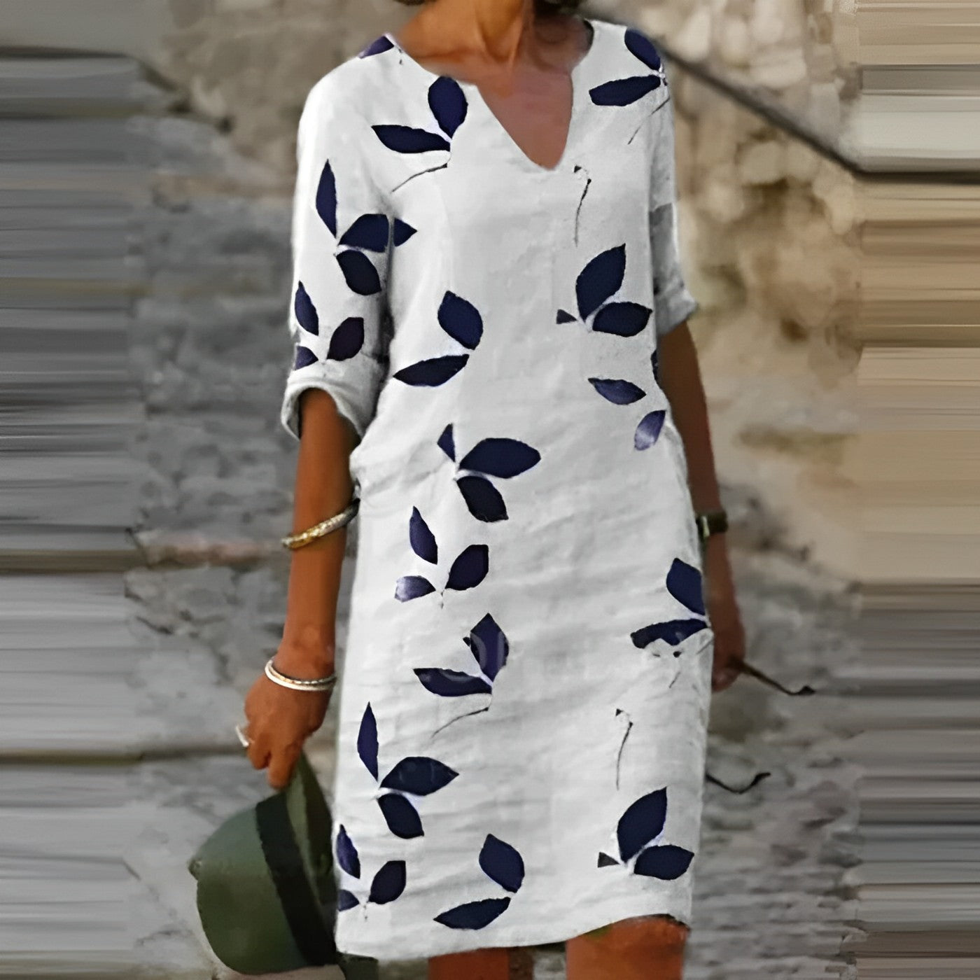 Abito estivo bianco con stampa foglie blu, maniche a tre quarti, scollo a V, indossato da donna su strada acciottolata. Moda casual elegante.