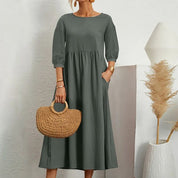 Abito lungo verde oliva con maniche a tre quarti, tasche laterali, borsa in paglia intrecciata, sfondo bianco minimalista, moda donna elegante.