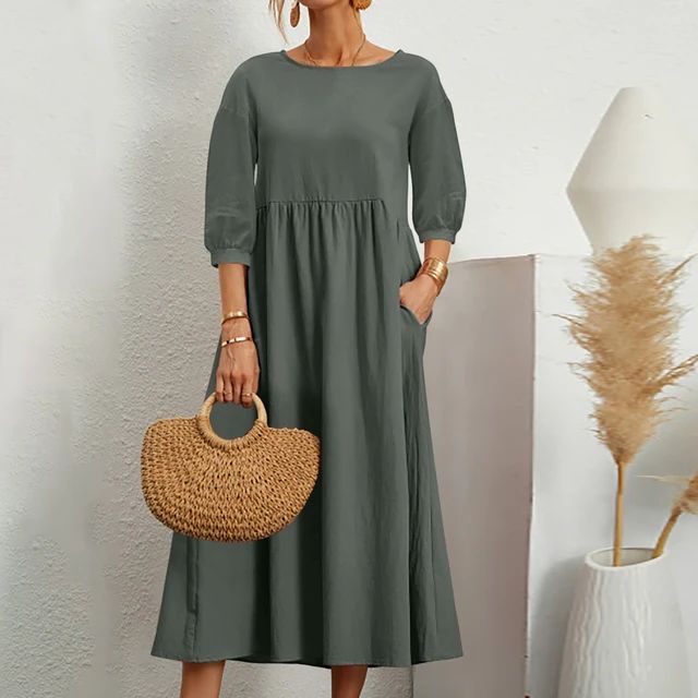 Abito lungo verde oliva con maniche a tre quarti, tasche laterali, borsa in paglia intrecciata, sfondo bianco minimalista, moda donna elegante.