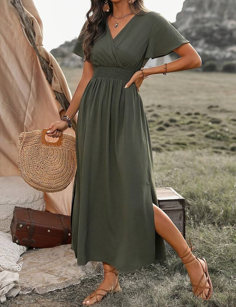 Donna in abito lungo verde oliva, sandali marroni, borsa di paglia, in un ambiente naturale con tenda e valigia. Moda estiva boho chic.