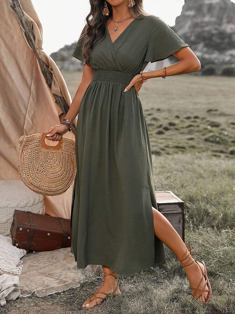 Donna in abito lungo verde oliva, sandali marroni, borsa di paglia, in un ambiente naturale con tenda e valigia. Moda estiva boho chic.