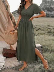 Donna in abito lungo verde oliva, sandali marroni, borsa di paglia, in un ambiente naturale con tenda e valigia. Moda estiva boho chic.