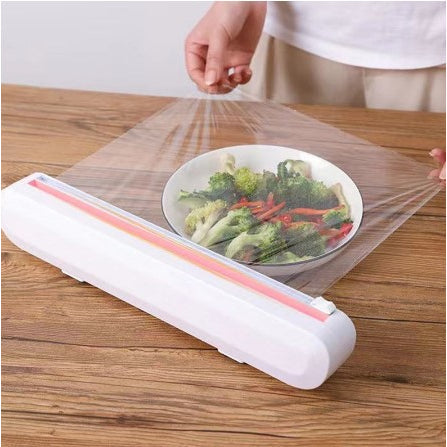 Dispenser pellicola trasparente su tavolo di legno, copre piatto con verdure fresche. Utensile cucina pratico, conservazione cibo efficace.