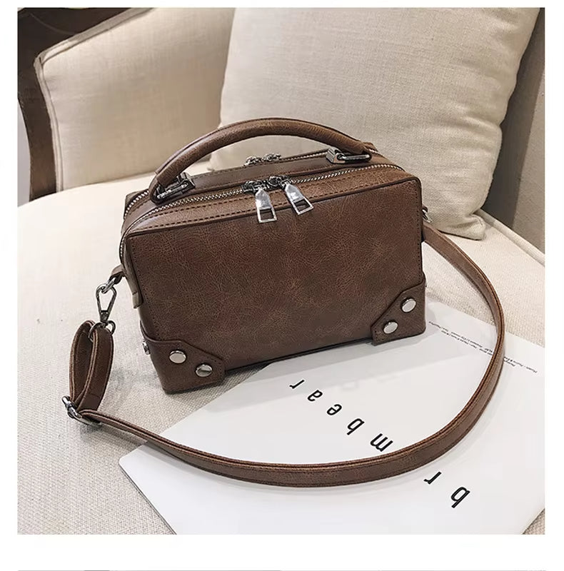 Borsa a tracolla in pelle marrone su sedia beige, design rettangolare con doppia zip e dettagli metallici, ideale per moda donna e accessori eleganti.