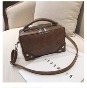Borsa a tracolla in pelle marrone su sedia beige, design rettangolare con doppia zip e dettagli metallici, ideale per moda donna e accessori eleganti.