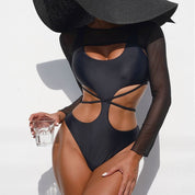 Donna in costume intero nero con dettagli cut-out e maniche lunghe trasparenti, indossa cappello a tesa larga, tiene bicchiere d'acqua.